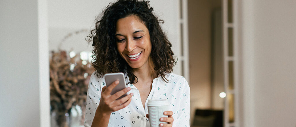 Woman in her home smiling, looking at her phone in one hand and holding a beverage in the other